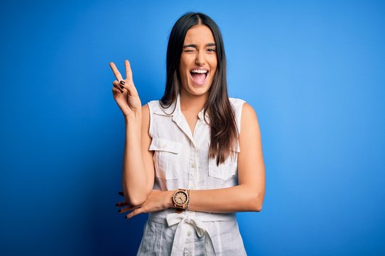 Young beautiful brunette woman wearing casual dress over isolated blue background smiling with happy face winking at the camera doing victory sign. Number two.