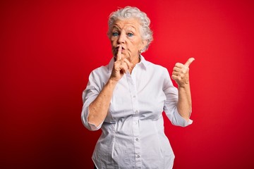 Senior beautiful woman wearing elegant shirt standing over isolated red background asking to be quiet with finger on lips pointing with hand to the side. Silence and secret concept.