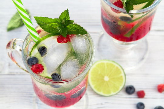 Homemade cold summer cocktail with mineral water, ice cubes, mint, lime, berry juice and fresh berries in glasses with straws