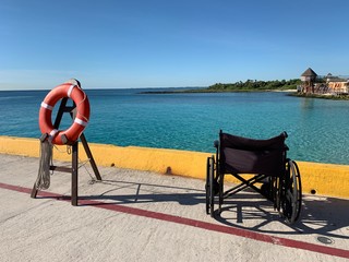 Black wheelchair is on the pier in Costa Maya, Mexico. Cruise ship guest left device. Red life ring  is ready to be used. Blue sea is on a background. View on a thatched roofs . - Powered by Adobe