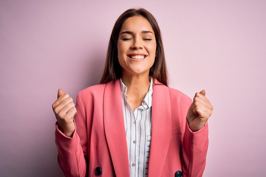 Young beautiful brunette woman wearing jacket standing over isolated pink background excited for success with arms raised and eyes closed celebrating victory smiling. Winner concept.