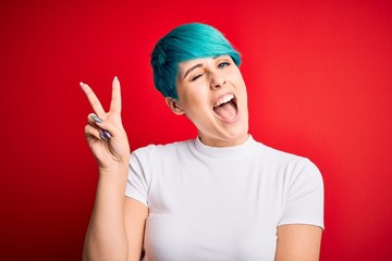 Young beautiful woman with blue fashion hair wearing casual t-shirt over red background smiling with happy face winking at the camera doing victory sign with fingers. Number two.