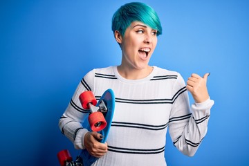 Young skater woman with blue fashion hair holding skateboard over blue isolated background pointing and showing with thumb up to the side with happy face smiling