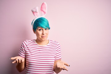 Young woman with fashion blue hair wearing easter rabbit ears over pink background clueless and...