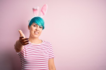 Young woman with fashion blue hair wearing easter rabbit ears over pink background smiling friendly offering handshake as greeting and welcoming. Successful business.