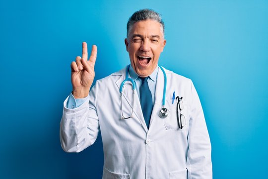 Middle age handsome grey-haired doctor man wearing coat and blue stethoscope smiling with happy face winking at the camera doing victory sign with fingers. Number two.