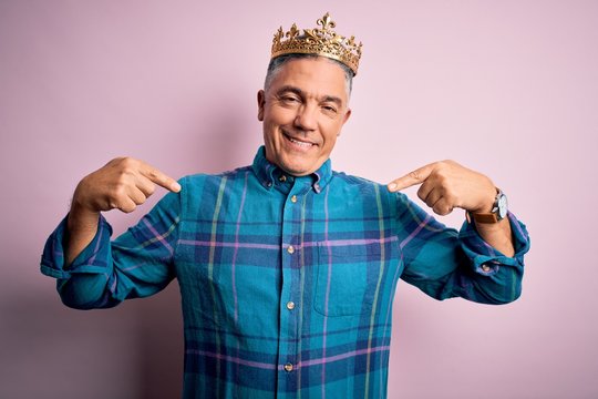 Middle age handsome grey-haired man wearing golden crown of king over pink background looking confident with smile on face, pointing oneself with fingers proud and happy.