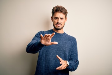 Young handsome man with beard wearing casual sweater standing over white background disgusted expression, displeased and fearful doing disgust face because aversion reaction. With hands raised