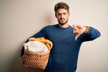 Young man doing housework holding wicker basket with clothes over white background with angry face,...