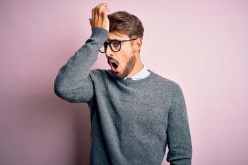 Young handsome man with beard wearing glasses and sweater standing over pink background surprised...