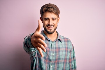 Young handsome man with beard wearing casual shirt standing over pink background smiling friendly offering handshake as greeting and welcoming. Successful business.