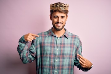 Young man with beard wearing golden crown of king standing over isolated pink background looking...