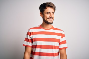 Fototapeta premium Young handsome man with beard wearing striped t-shirt standing over white background looking away to side with smile on face, natural expression. Laughing confident.