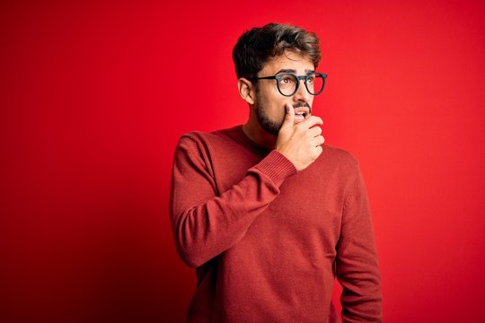 Young handsome man with beard wearing glasses and sweater standing over red background Thinking worried about a question, concerned and nervous with hand on chin