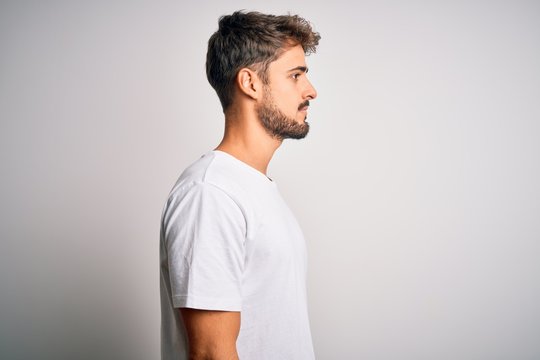 Young handsome man with beard wearing casual t-shirt standing over white background looking to side, relax profile pose with natural face with confident smile.