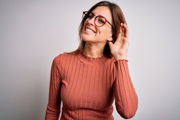 Young beautiful brunette woman wearing casual sweater and glasses over white background smiling with hand over ear listening an hearing to rumor or gossip. Deafness concept.