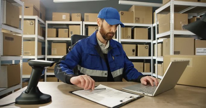 Caucasian young man in uniform and cap working at laptop computer in post office store with parcels. Postman registering mail parcel it and filling in invoice document in postal storage room.