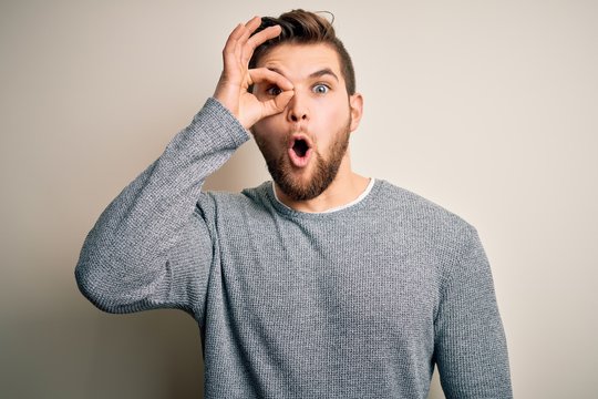 Young handsome blond man with beard and blue eyes wearing casual sweater doing ok gesture shocked with surprised face, eye looking through fingers. Unbelieving expression.