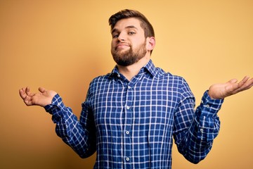 Young blond businessman with beard and blue eyes wearing shirt over yellow background clueless and confused expression with arms and hands raised. Doubt concept.