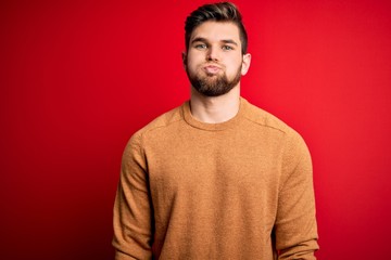 Young blond man with beard and blue eyes wearing casual sweate over red background puffing cheeks with funny face. Mouth inflated with air, crazy expression.