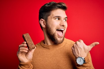 Young blond man with beard and blue eyes eating healthy chocolate protein bar pointing and showing...