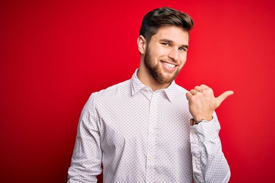 Young blond businessman with beard and blue eyes wearing elegant shirt over red background smiling with happy face looking and pointing to the side with thumb up.
