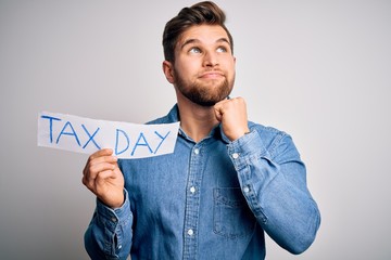 Young blond man with beard and blue eyes holding paper with tax day message serious face thinking...