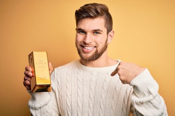 Young blond man with beard and blue eyes holding gold ingot over isolated yellow background with...