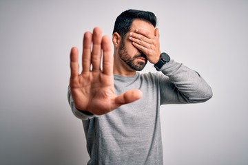 Young handsome man with beard wearing casual sweater standing over white background covering eyes with hands and doing stop gesture with sad and fear expression. Embarrassed and negative concept.