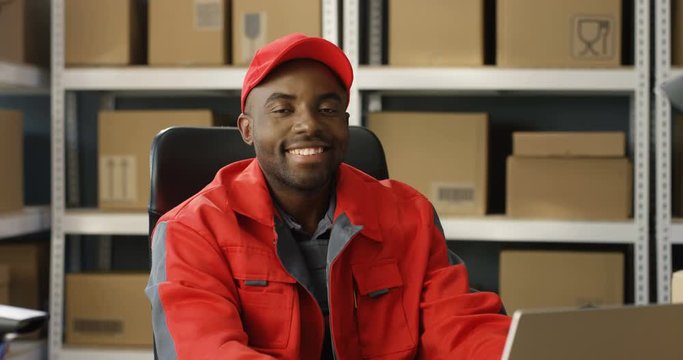 Young cheerful African American postman in red iniform and cap sitting at desk in postal office store and working at computer. Portrait of male courier smiling happily to camera in storage of parcels.