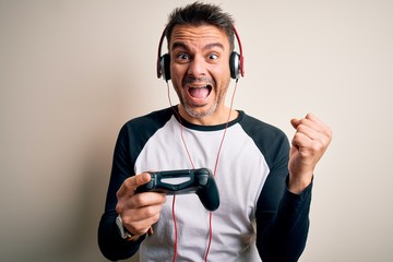 Young handsome gamer man playing video game using joystick and headphones screaming proud and celebrating victory and success very excited, cheering emotion