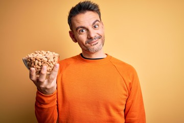 Young handsome man holding bowl with peanuts standing over isolated yellow background with a happy face standing and smiling with a confident smile showing teeth