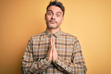 Young handsome man wearing casual shirt standing over isolated yellow background praying with hands together asking for forgiveness smiling confident.