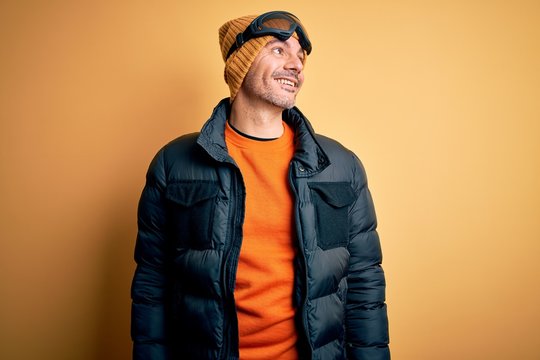 Young handsome skier man skiing wearing snow sportswear using ski goggles looking away to side with smile on face, natural expression. Laughing confident.