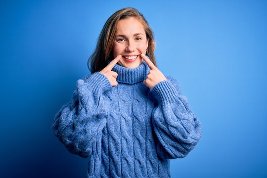 Young beautiful blonde woman wearing casual turtleneck sweater over blue background Smiling with open mouth, fingers pointing and forcing cheerful smile