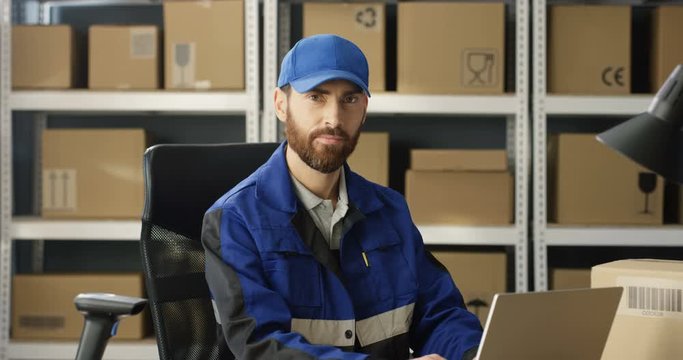 Young joyful Caucasian postman in iniform and cap sitting at desk in postal office store and working at computer. Man typing on laptop. Portrait of male courier smiling to camera in storage of boxes.