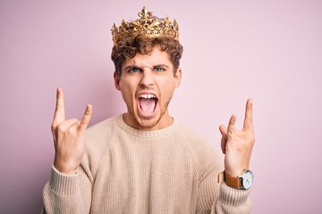 Young blond man with curly hair wearing golden crown of king over pink background shouting with...