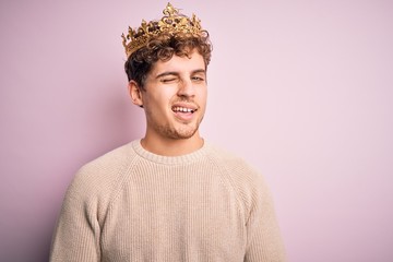 Young blond man with curly hair wearing golden crown of king over pink background winking looking...