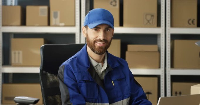 Portrait of young happy Caucasian postman in iniform and cap sitting at desk in postal office store and working at computer. Handsome male courier smiling happily to camera in storage of parcels.