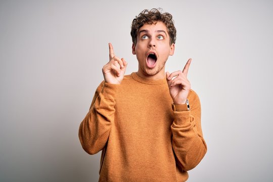 Young blond handsome man with curly hair wearing casual sweater over white background amazed and surprised looking up and pointing with fingers and raised arms.
