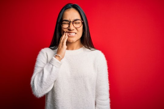 Young beautiful asian woman wearing casual sweater and glasses over red background touching mouth with hand with painful expression because of toothache or dental illness on teeth. Dentist