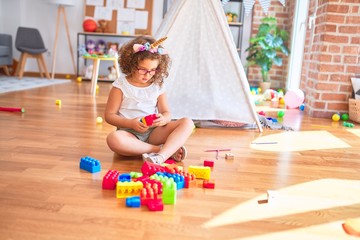 Beautiful toddler wearing glasses and unicorn diadem sitting playing with building blocks at kindergarten