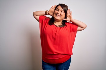 Beautiful brunette plus size woman wearing casual red t-shirt over isolated white background Smiling cheerful playing peek a boo with hands showing face. Surprised and exited