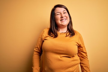 Beautiful brunette plus size woman wearing casual sweater over isolated yellow background winking looking at the camera with sexy expression, cheerful and happy face.