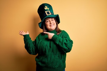 Young beautiful plus size woman wearing green hat with clover celebrating saint patricks day amazed and smiling to the camera while presenting with hand and pointing with finger.