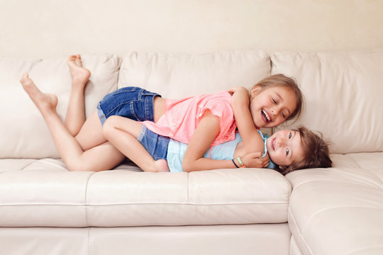 Two cute little Caucasian girls siblings playing at home. Adorable children kids lying hugging on a couch together. Authentic candid lifestyle domestic life moment. Happy friends sisters relationship.