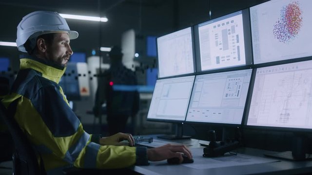 Industry 4.0 Modern Factory: Facility Operator Controls Workshop Production Line, Uses Computer with Screens Showing Complex UI of Machine Operation Processes, Controllers, Machinery Blueprints