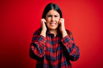 Young beautiful woman wearing casual shirt over red background covering ears with fingers with annoyed expression for the noise of loud music. Deaf concept.