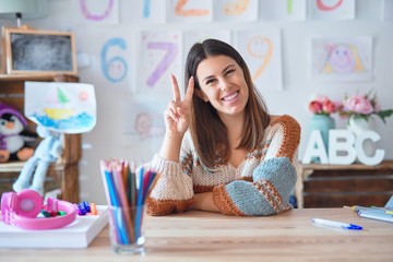 Young beautiful teacher woman wearing sweater and glasses sitting on desk at kindergarten smiling...