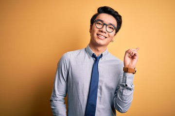 Young handsome chinese businessman wearing glasses and tie over yellow background with a big smile on face, pointing with hand and finger to the side looking at the camera.